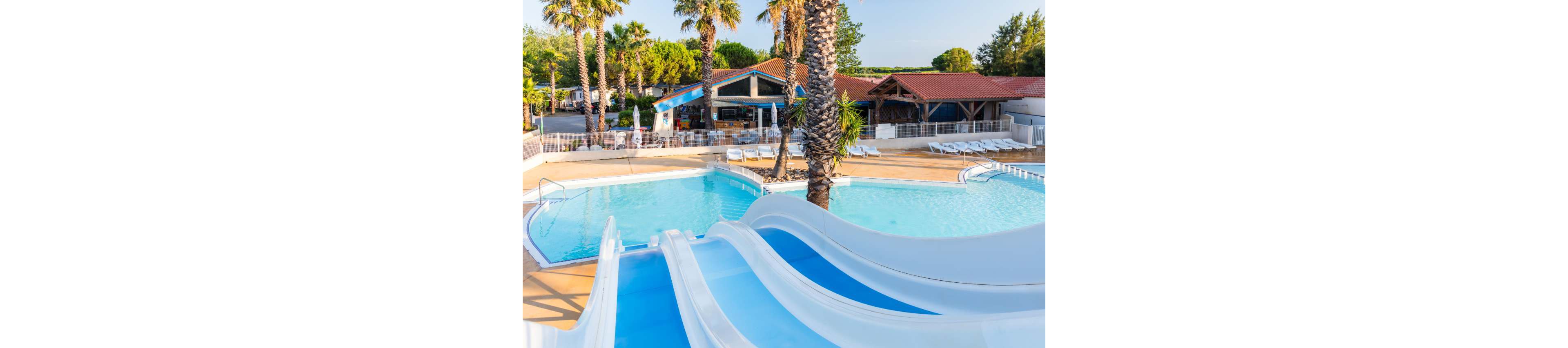 A bright outdoor resort pool features multiple white and blue waterslides leading into clear turquoise water. Palm trees and lush greenery surround the pool area, creating a tropical vacation atmosphere. In the background, a poolside bar or café with red-tiled roofs and shaded seating is visible. White lounge chairs line the deck, suggesting a relaxed and family-friendly holiday setting.