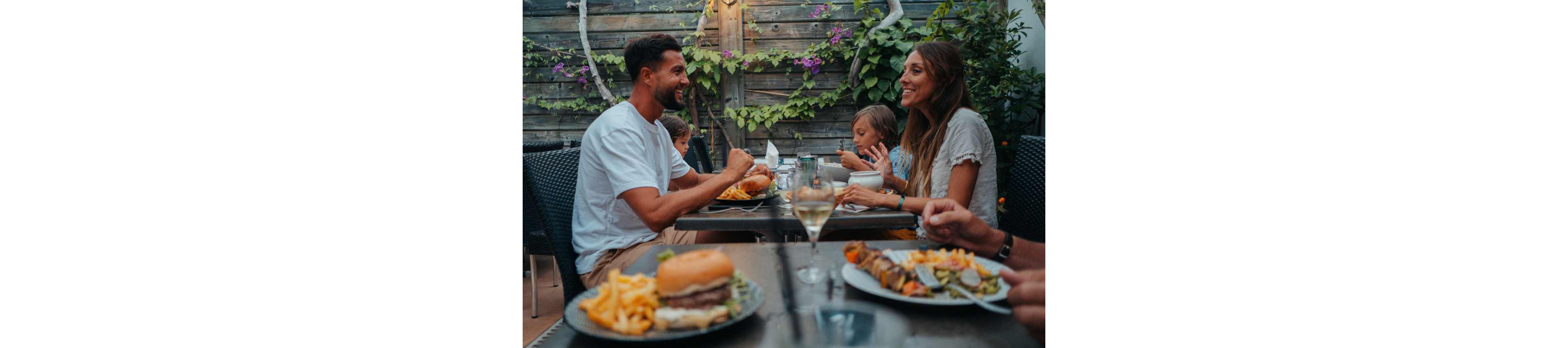 A family sits together at an outdoor restaurant table sharing a meal. Plates of food including burgers, fries, and skewers are arranged in front of them. A glass of white wine and other drinks are visible on the table. The setting features a wooden fence with green vines and purple flowers, creating a relaxed, casual atmosphere. The scene suggests a warm evening gathering focused on conversation and dining.