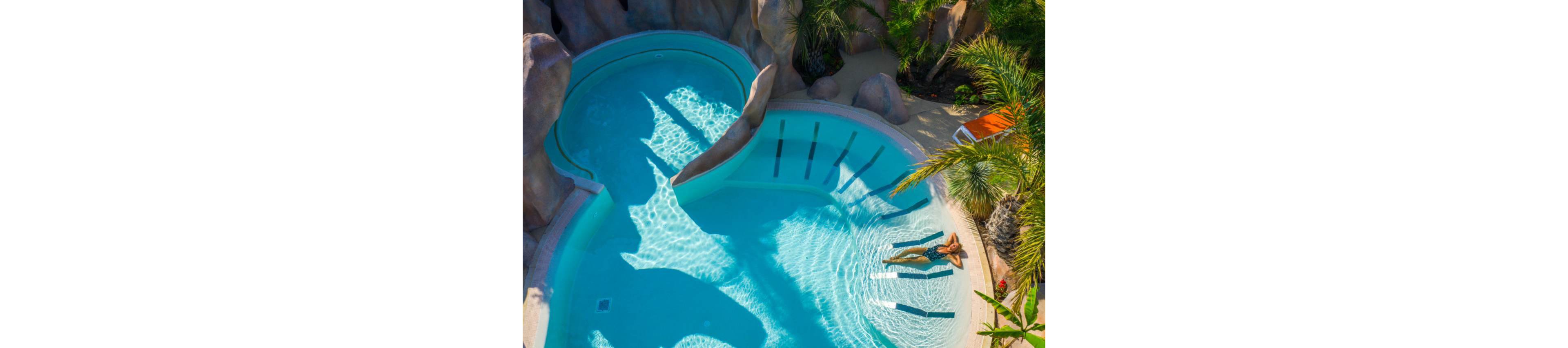 An aerial view shows a woman floating on a lounger in a bright blue swimming pool. The pool features curved shapes, rock-like edges, and a connected circular spa area. Sunlight creates shimmering patterns on the clear water, emphasizing the tranquil atmosphere. Lush palm trees and a nearby lounge chair suggest a tropical resort setting.