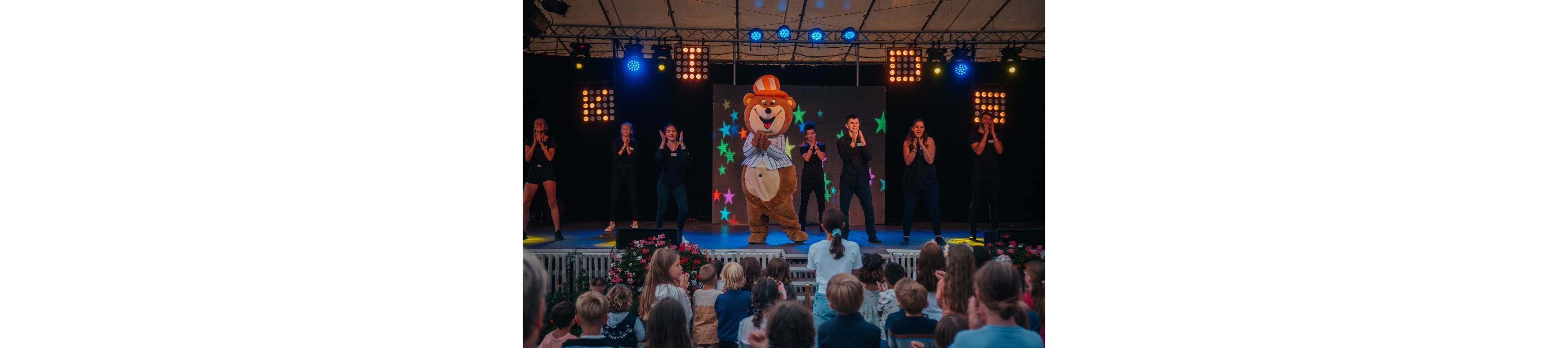A large bear mascot in a striped shirt and orange hat performs on a brightly lit stage. Several dancers dressed in dark outfits stand on either side, clapping and dancing in unison. Colorful star graphics glow on a screen behind them, with multiple stage lights illuminating the scene. A crowd of children and adults watch from the foreground, creating a lively family entertainment atmosphere.