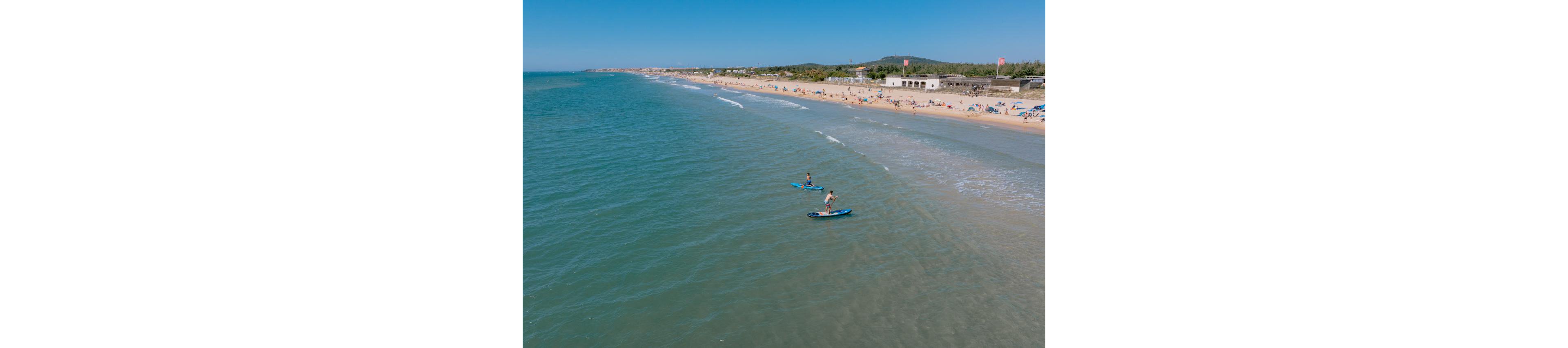 Two people on stand up paddleboards glide over calm coastal waters near a wide sandy beach. The shoreline is dotted with sunbathers and umbrellas under a clear blue sky. Gentle waves roll toward the shore, creating a relaxed seaside atmosphere. Low buildings and distant hills frame the background, emphasizing the expansive coastline.