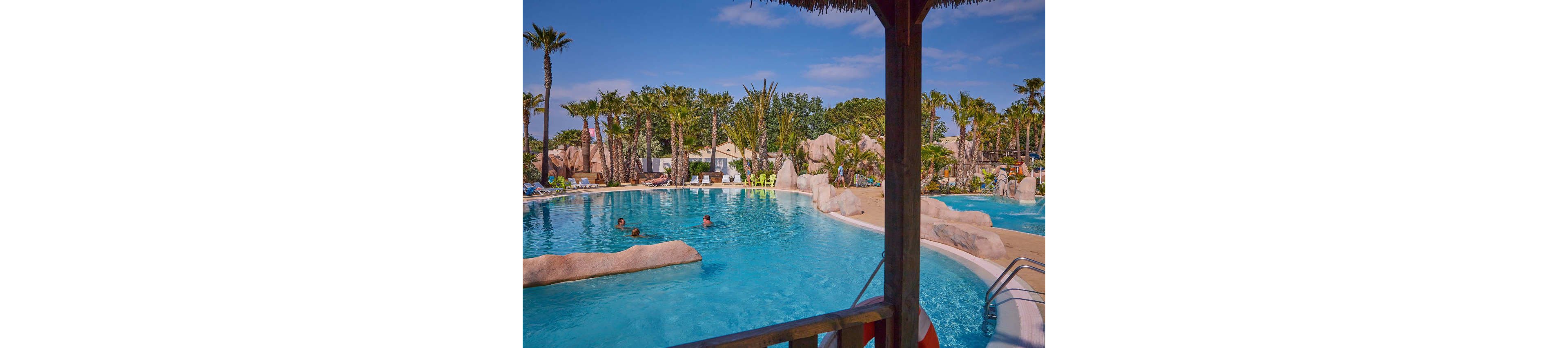 A large resort-style swimming pool is surrounded by numerous palm trees and rock formations, creating a tropical oasis atmosphere. Several people are swimming and relaxing in the clear blue water. Sun loungers and colorful chairs line the far side of the pool, suggesting a vacation or holiday setting. The scene is viewed from a shaded wooden structure, with a life preserver partially visible in the foreground.