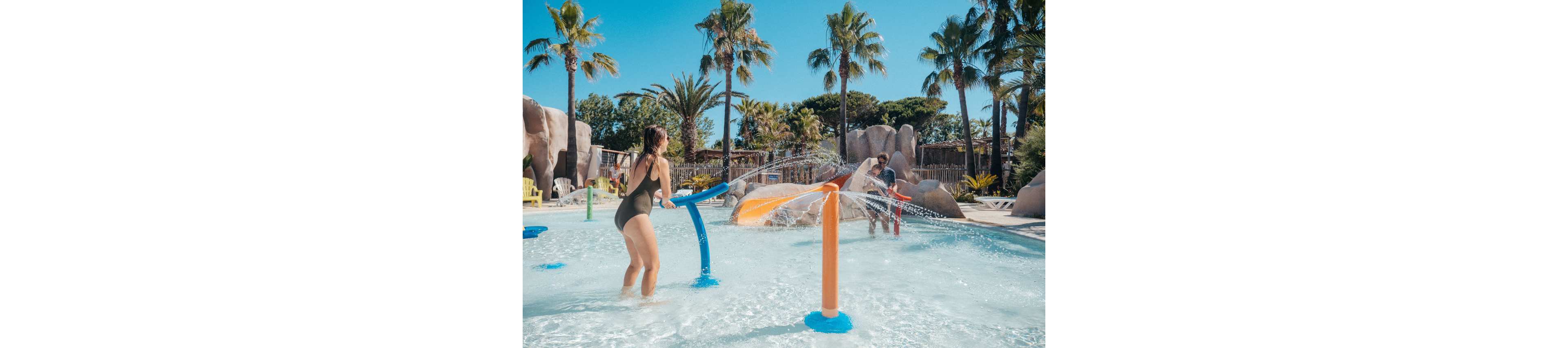 A young woman in a swimsuit plays with a water sprayer in a shallow splash pool. In the background, children enjoy a small orange slide and other colorful water features. Palm trees, rocks, and clear blue sky create a tropical resort atmosphere. The scene captures a playful summer day at an outdoor water park.