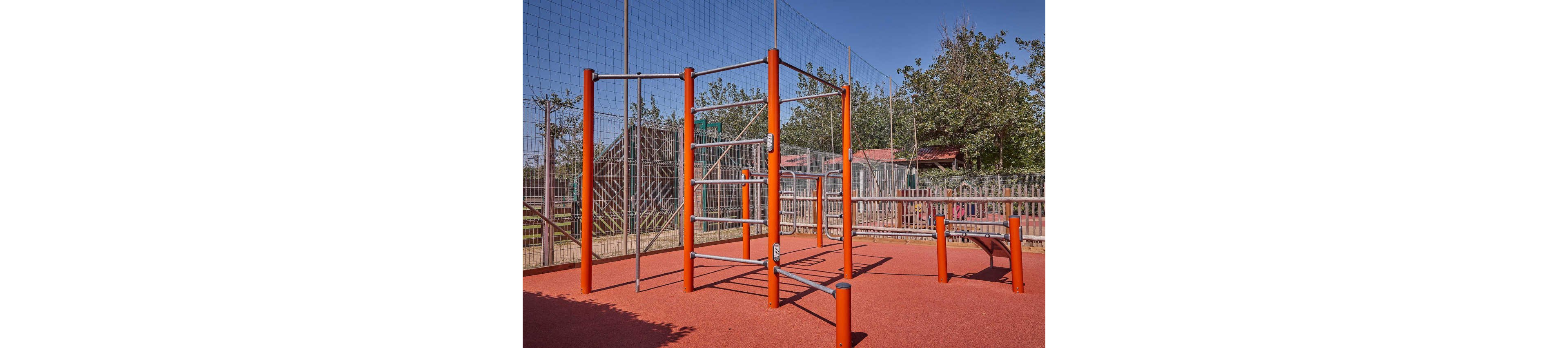 An outdoor fitness area features bright red metal exercise structures on a rubberized surface. Parallel bars, ladders, and horizontal bars are arranged within a fenced playground. Trees and small buildings are visible in the background under a clear blue sky. The space appears designed for bodyweight workouts and recreational exercise.