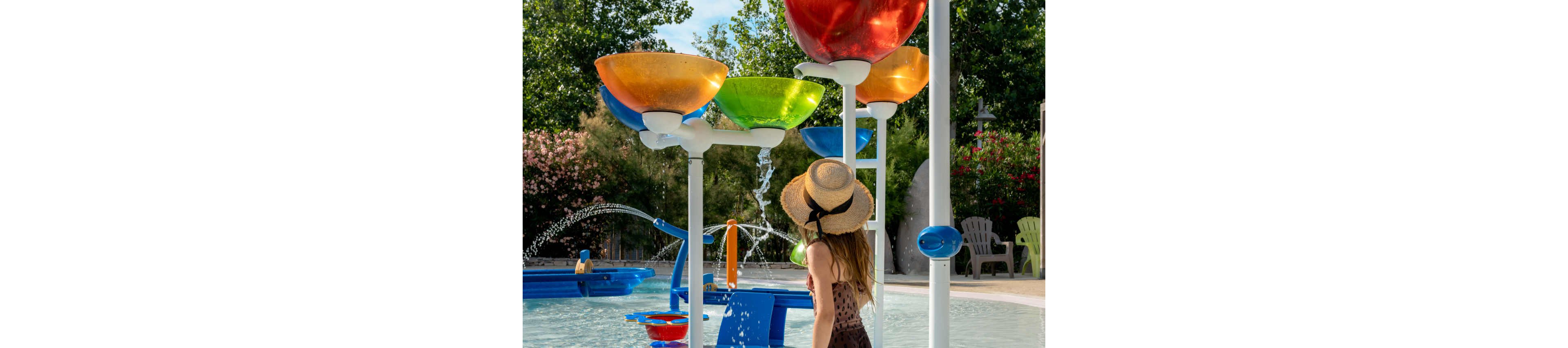 A young girl in a straw hat stands at a splash pad, watching large colorful bowls that pour water into a shallow pool. The scene is set in an outdoor water play area with fountains spraying in the background. Bright red, orange, green, and blue structures create a playful, vibrant atmosphere. Trees, flowers, and patio chairs surround the area, suggesting a family-friendly recreational space.