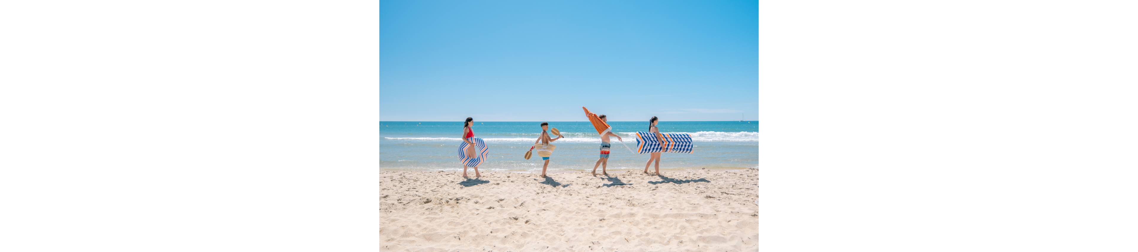 Four young friends walk in a line along a sunny sandy beach, each carrying colorful inflatable floats. The clear blue ocean and gentle waves create a bright, summery backdrop under a cloudless sky. Two people hold large striped tube floats while another carries an orange bodyboard-style float. The scene captures a carefree vacation moment as they head toward the water for fun.