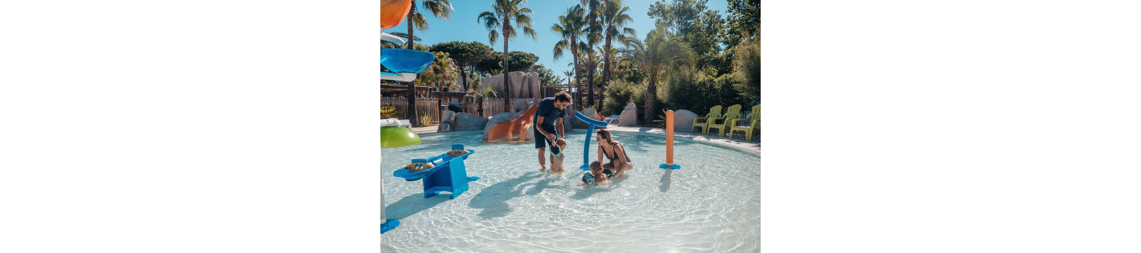 A family with two young children plays together in a shallow outdoor splash pool. The parents help the toddlers explore the water features and small slides under a bright sunny sky. Palm trees, colorful play structures, and lounge chairs surround the pool, creating a relaxed vacation atmosphere. The scene feels warm, joyful, and family friendly.