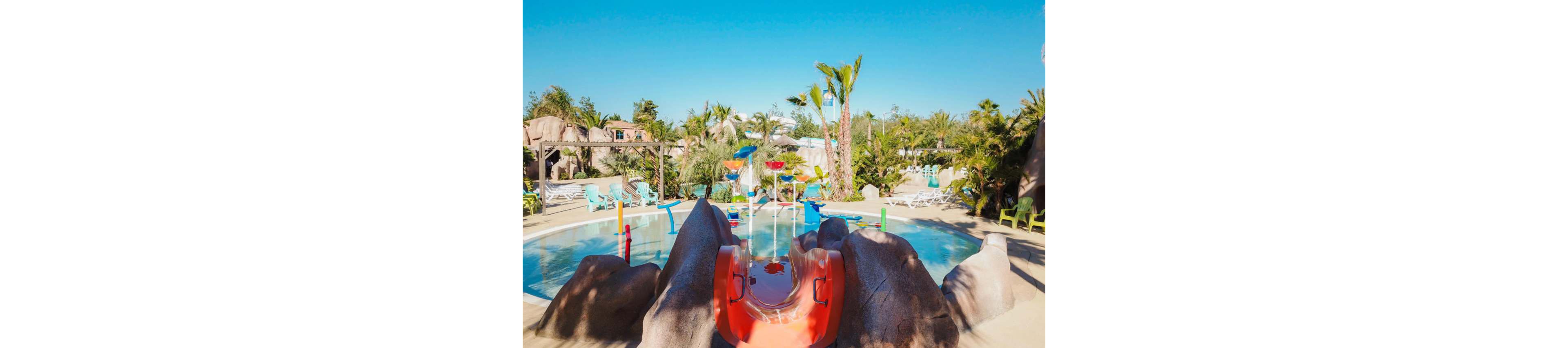 A bright children’s splash pool features a small red slide framed by artificial rocks. Colorful water play structures and fountains are set in the shallow circular pool. Palm trees and lush greenery surround the area, creating a tropical resort atmosphere. Sun loungers and chairs are arranged around the pool under a clear blue sky, suggesting a sunny vacation setting.