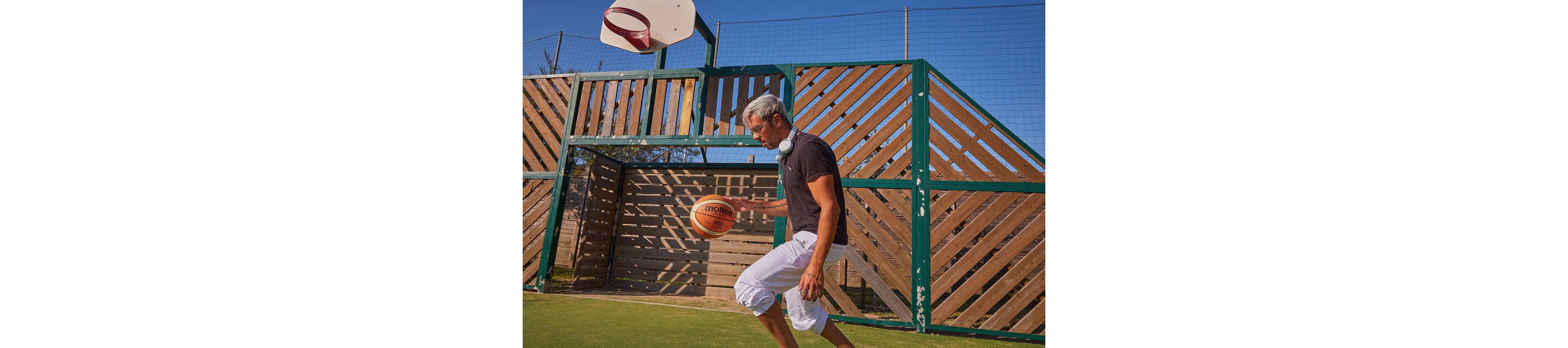 A young man dribbles a basketball on an outdoor court. He wears casual clothing and headphones around his neck while moving toward the hoop. The court features a wooden and metal fence backdrop under a clear blue sky. The scene suggests an informal game or solo basketball practice.
