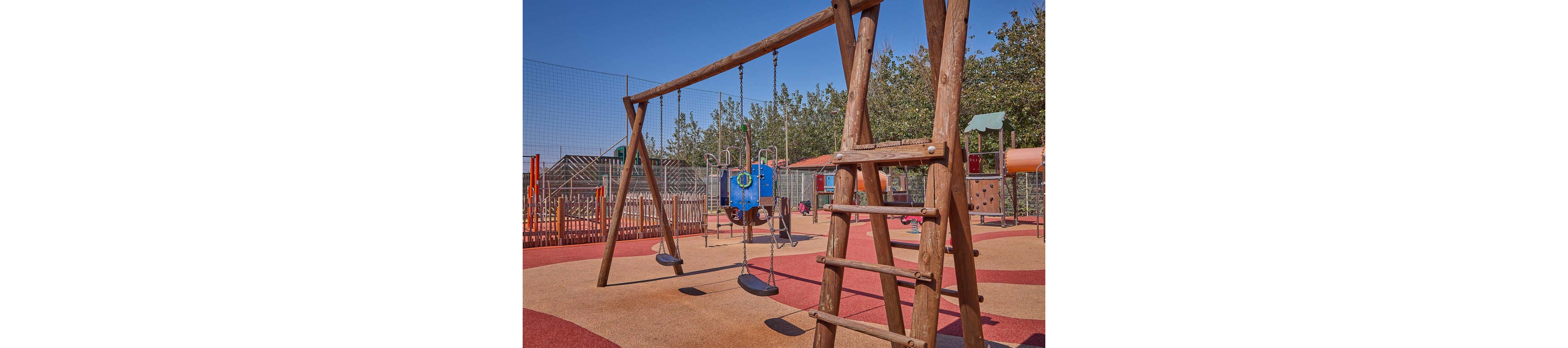 A wooden swing set with two seats stands in the foreground of an empty playground. The play area features colorful rubber flooring in red and orange tones and a climbing structure with a slide in the background. Trees and a protective fence surround the playground under a clear blue sky. The scene appears calm and well-maintained, suggesting a safe outdoor space for children.