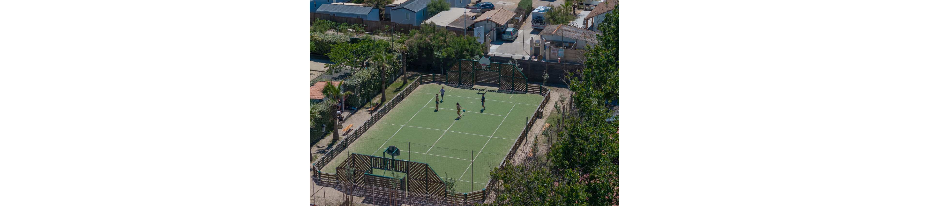 An aerial view shows a small group of people playing on an outdoor tennis court. The green court is enclosed by wooden fencing and surrounded by trees and shrubs. Nearby, residential buildings and parked vehicles indicate a neighborhood or vacation setting. The bright daylight and clear weather create a relaxed, recreational atmosphere.