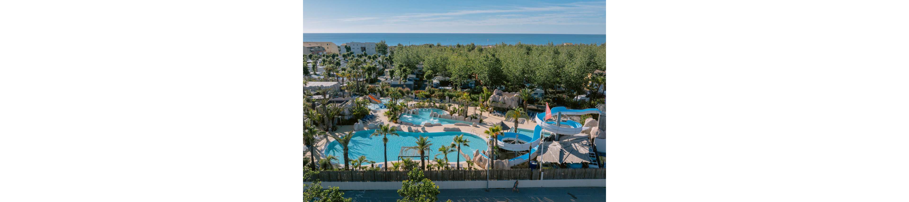 An aerial view shows a large outdoor water park with multiple pools and winding water slides surrounded by palm trees. The complex is set near a coastline, with the sea visible in the background beyond a dense line of trees. Curved pools, sun loungers, and landscaped pathways create a resort-like atmosphere. The scene suggests a sunny vacation destination focused on leisure and family fun.
