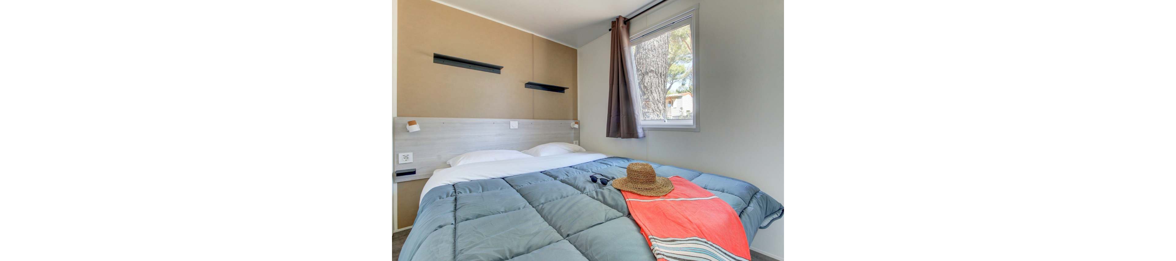 A neatly made double bed with a blue comforter is the main focus of this small bedroom. A straw sun hat and a colorful striped towel are placed on the bed, suggesting a relaxed vacation setting. The room features neutral walls, a window with curtains letting in daylight, and simple modern shelves above the headboard. Overall, the scene feels bright, tidy, and inviting.