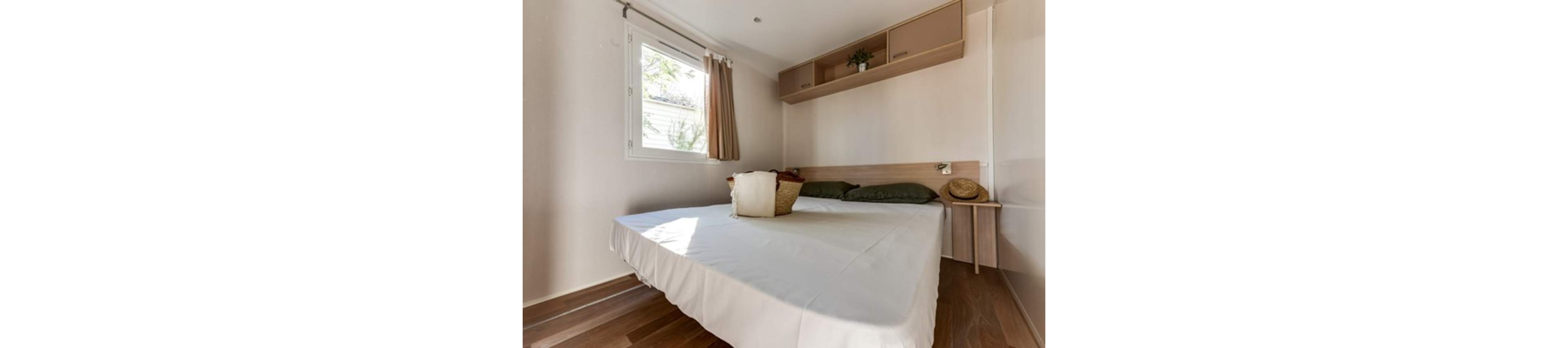 A compact bedroom features a neatly made double bed with a white sheet and pillows. Natural light enters through a window with beige curtains on the left wall. Above the bed, open shelving holds a small potted plant and decor items, adding a touch of greenery. A small bedside table on the right holds a woven hat, while a basket sits on the bed, enhancing the cozy, minimalist atmosphere.
