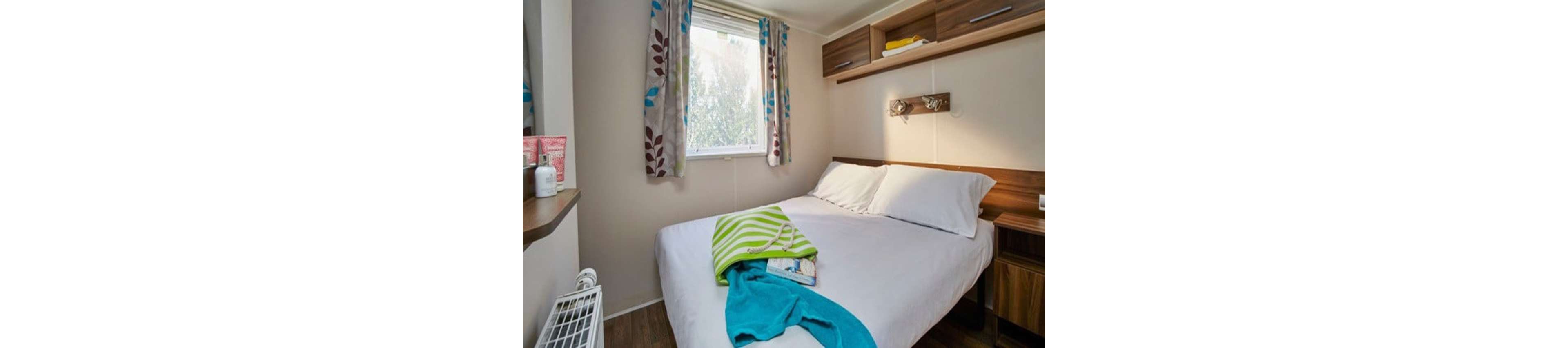 A small, cozy bedroom features a neatly made double bed with white linens. A bright window with patterned curtains lets in natural light, illuminating the compact space. Colorful towels and a magazine rest on the bed, adding a casual, lived-in feel. Wooden shelves, a wall-mounted light, and a small heater complete the simple, functional decor.