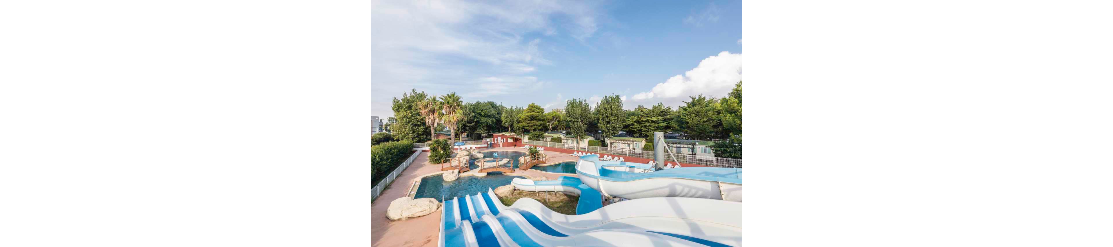 An outdoor water park features multiple curved white and blue water slides leading into clear pools. The area is empty, with rows of white lounge chairs arranged around the pool deck. Green trees and palm trees surround the park, with a fence and parked cars visible in the background. The sky is bright with scattered clouds, suggesting a sunny, pleasant day. The overall scene conveys a clean, family-friendly recreational environment.