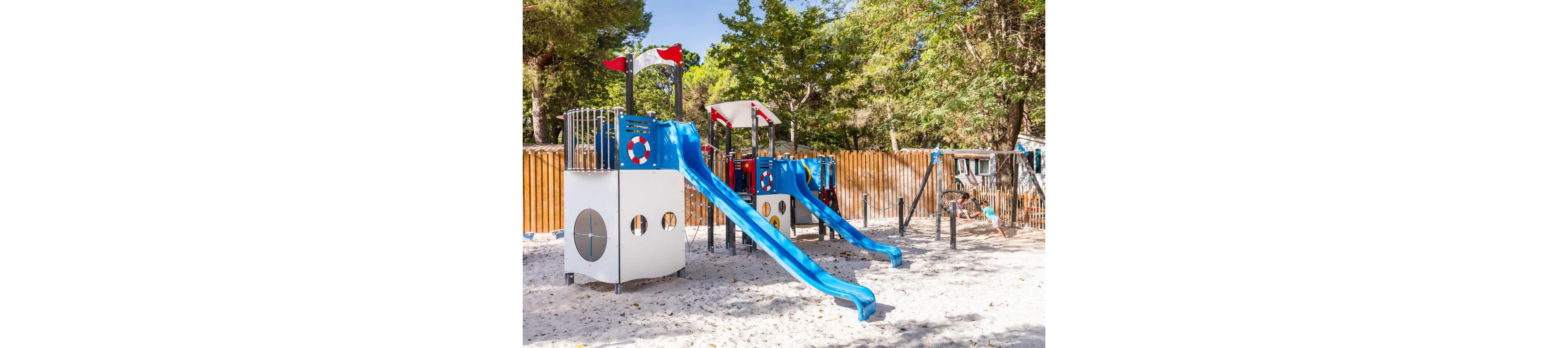 A brightly colored playground structure with two blue slides stands on a sandy surface. The play equipment features nautical-themed details such as lifebuoy designs and circular windows. A wooden fence and tall green trees surround the area, creating a shaded outdoor setting. A child plays in the background, adding a sense of activity and fun to the scene.