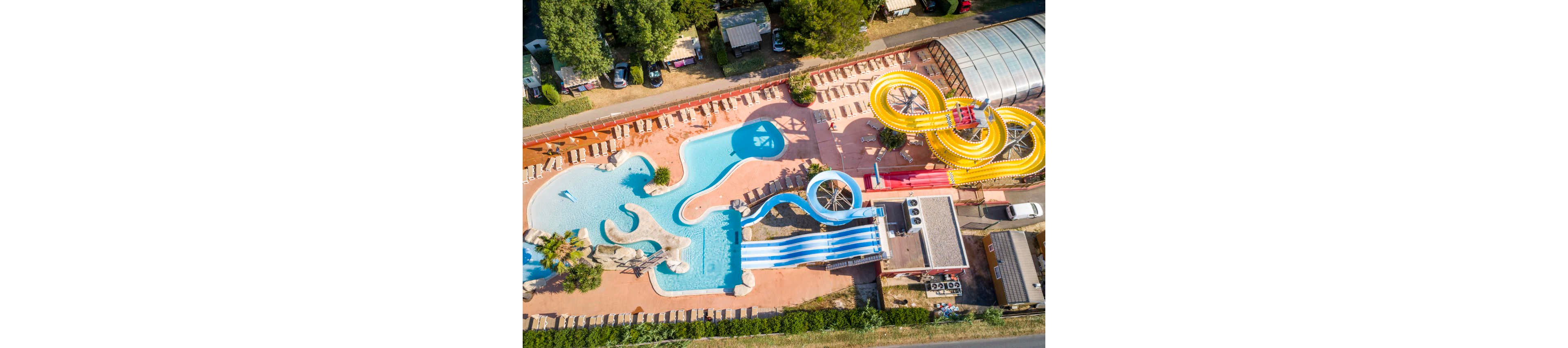 A bright outdoor water park is captured from above, showing winding pools and multiple slides. The main pool area features curving blue and white slides that end in shallow water. On the right side, a large yellow spiral slide structure stands next to a covered section. Sun loungers line the perimeter, and surrounding trees and buildings frame the recreational area. The scene suggests a sunny, family-friendly leisure destination.