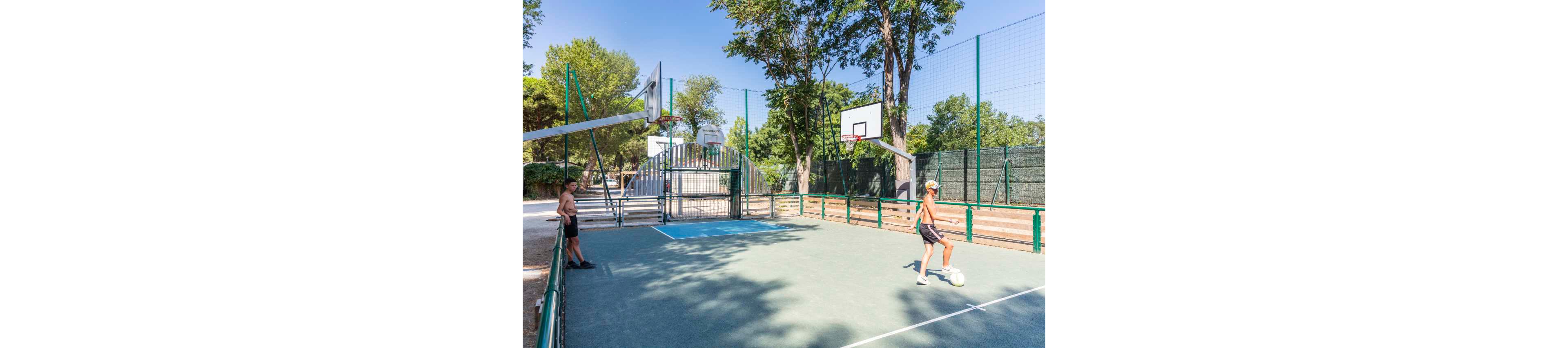 Two teenagers are on a multi-sport outdoor court, with one dribbling a soccer ball while the other stands near the sideline. The court features basketball hoops, a fenced perimeter, and a small grandstand area in the background. Tall trees and greenery surround the court, creating a bright, sunny park setting. The scene suggests casual recreation and summer leisure in a safe, open environment.