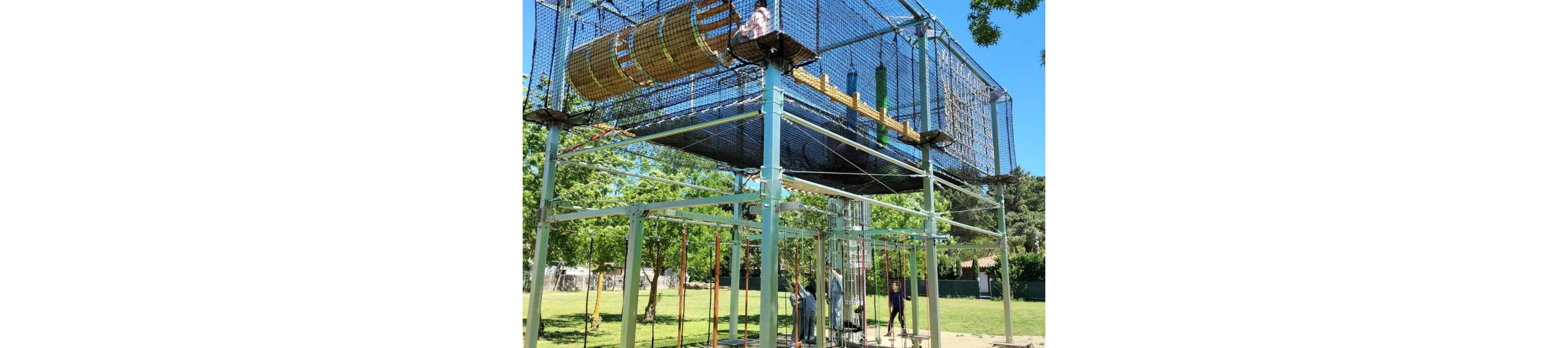An elevated rope course structure stands in a sunny park, with children climbing and exploring different levels. The course features netted walkways, hanging ropes, and cylindrical tunnels made of wood and mesh. Adults and kids gather below the structure, preparing to climb or watching from the ground. Lush green trees and a bright blue sky create a cheerful outdoor atmosphere.