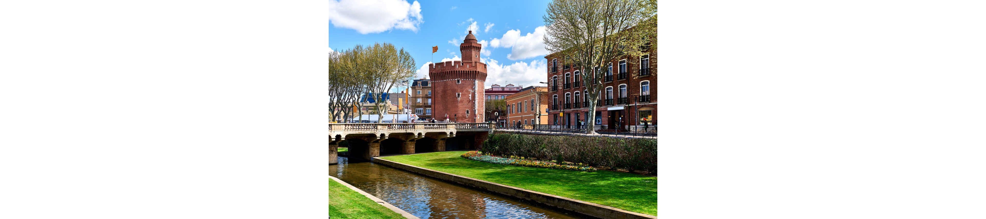 A historic red brick tower rises prominently beside a calm canal in a European city. A stone bridge spans the waterway, connecting green lawns and manicured flowerbeds on either side. Leafy trees line the promenade, framing the scene against a bright blue sky with scattered clouds. Traditional multi-story buildings border the street, adding to the old-town atmosphere.
