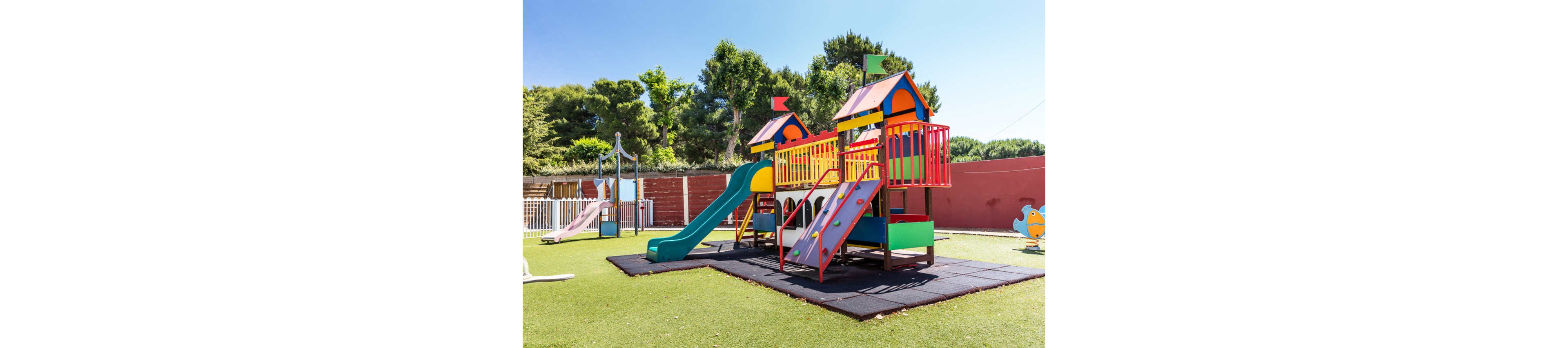 A bright, colorful playground stands in an outdoor area under a clear blue sky. The main structure features two small towers connected by platforms, with red railings and multicolored panels. A green slide, a red climbing ramp, and other play elements sit on safety mats over artificial grass. Trees and a red fence enclose the play area, creating a safe and cheerful environment for children. The scene is empty of people, emphasizing the equipment and sunny weather.
