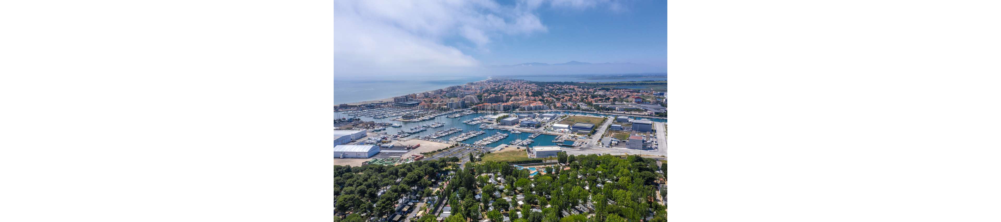 An aerial view captures a large coastal marina filled with numerous boats and yachts. The harbor is surrounded by industrial buildings, residential neighborhoods, and a dense green area in the foreground. Beyond the town, the shoreline stretches along a calm sea under a partly cloudy sky. Distant hills or mountains are faintly visible on the horizon, adding depth to the coastal landscape.