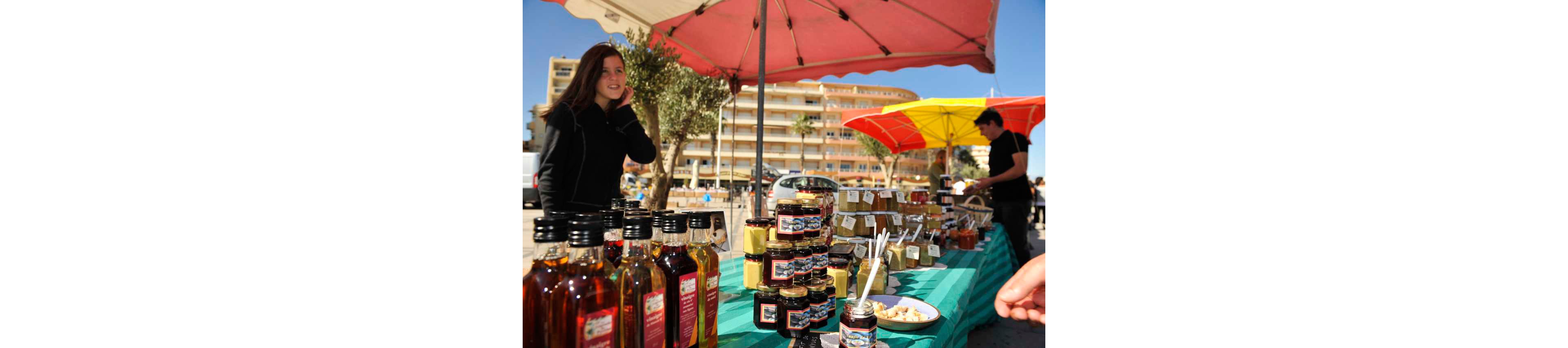 An outdoor market stall displays an assortment of bottled oils and jars of preserves on a green tablecloth. The scene is set in a sunny urban square with apartment buildings and palm trees in the background. A woman walks past the stall while another person stands behind the table under colorful umbrellas. Small labels are attached to many of the jars, and tasting spoons are placed in a sample jar at the front. The bright daylight and vivid umbrella colors create a lively, inviting atmosphere.