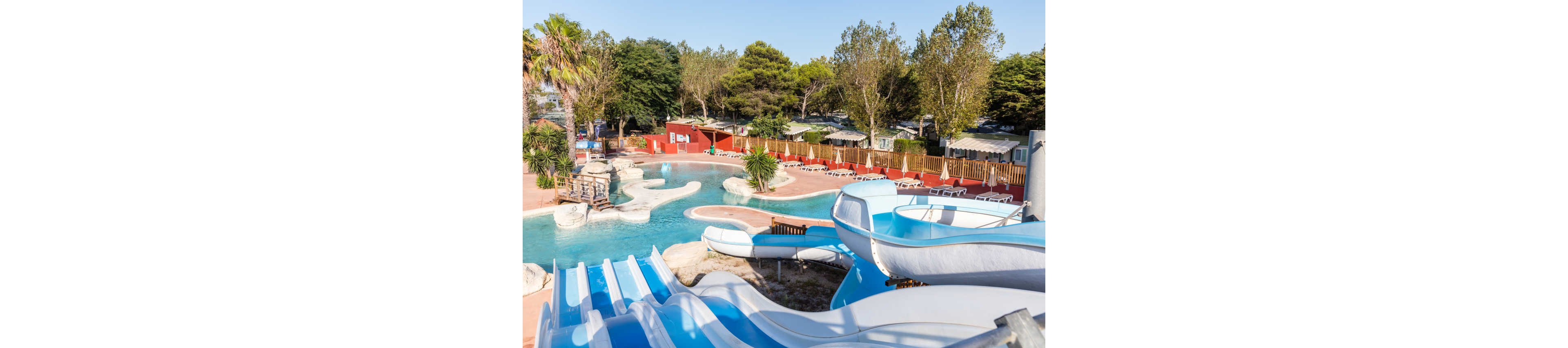 An outdoor water park features multiple blue and white water slides leading into a winding pool. The setting includes sun loungers arranged around the poolside, suggesting a relaxing vacation atmosphere. Trees and greenery surround the area, with cabins or mobile homes visible in the background behind a wooden fence. The bright sunlight and clear sky create a cheerful, family-friendly resort scene.