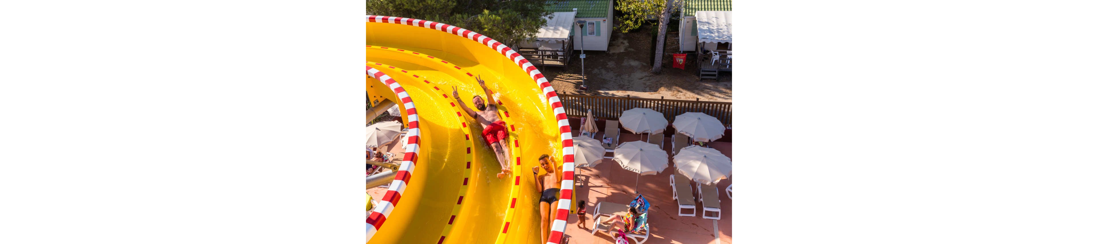 An overhead view shows two people sliding down a large yellow water slide with red accents. The slide curves through a sunny outdoor recreation area near holiday cabins and trees. Below, several white umbrellas shade lounge chairs where people relax and watch. The scene captures a fun summer atmosphere at a resort or campground pool area.