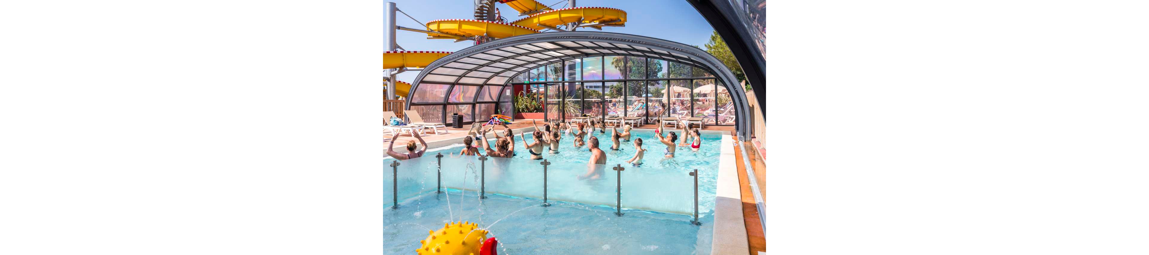 A group of adults participates in an aqua aerobics class in a shallow, covered swimming pool. The transparent arched roof shelters the pool while allowing sunlight and views of the surrounding resort area. In the background, people relax on sun loungers and a large yellow water slide winds above the scene. The atmosphere is lively and summery, with bright blue water and colorful structures creating a fun vacation setting.