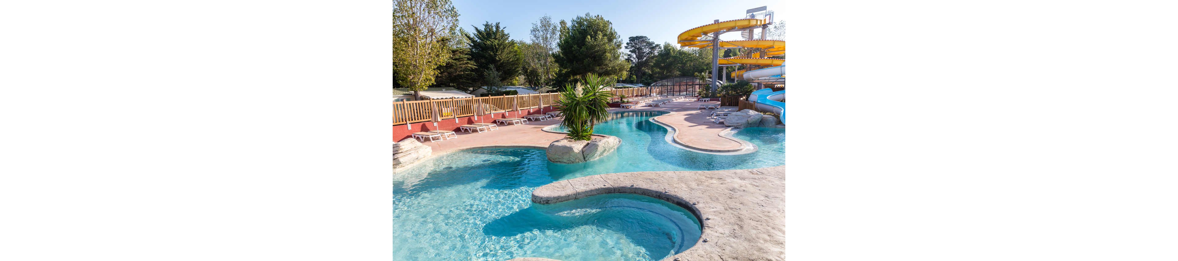An outdoor water park features a winding lazy river surrounded by smooth stone edges and clear blue water. A small island with tropical plants sits in the center of the pool, adding a resort-like feel. In the background, yellow water slides curve above the complex and lounge chairs line the deck behind a wooden fence. The scene is bright and sunny, suggesting a warm day perfect for swimming and relaxation.