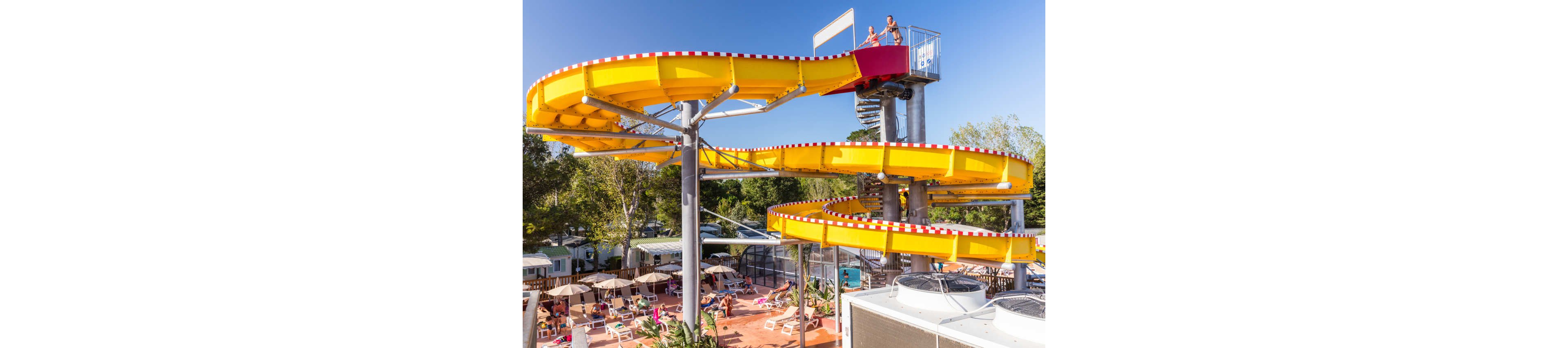 A large yellow waterslide spirals above an outdoor resort area filled with sunbathers and swimmers. People relax on lounge chairs under umbrellas while others enjoy the pool below. The slide structure features a tall metal staircase and bright red-and-white edging, standing out against the surrounding trees and cabins. The sunny weather and vibrant colors create a lively vacation atmosphere.