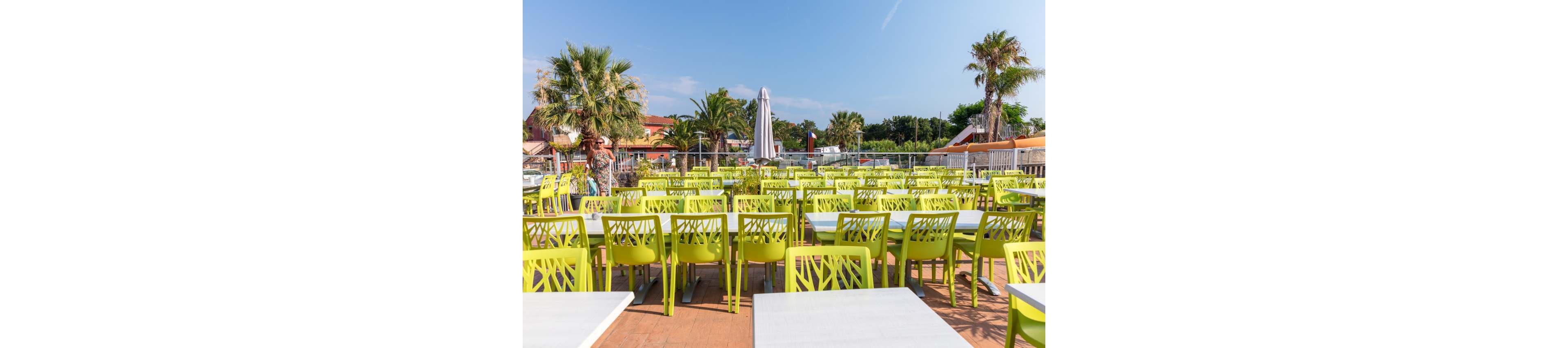 An outdoor swimming pool is filled with people swimming and relaxing under a clear blue sky. The long rectangular pool has lane dividers and starting blocks, suggesting a space for both leisure and lap swimming. Sunbathers lounge on chairs along the poolside terrace near a low modern building with balconies and palm trees. The overall scene conveys a warm, sunny vacation atmosphere at a resort or campground.