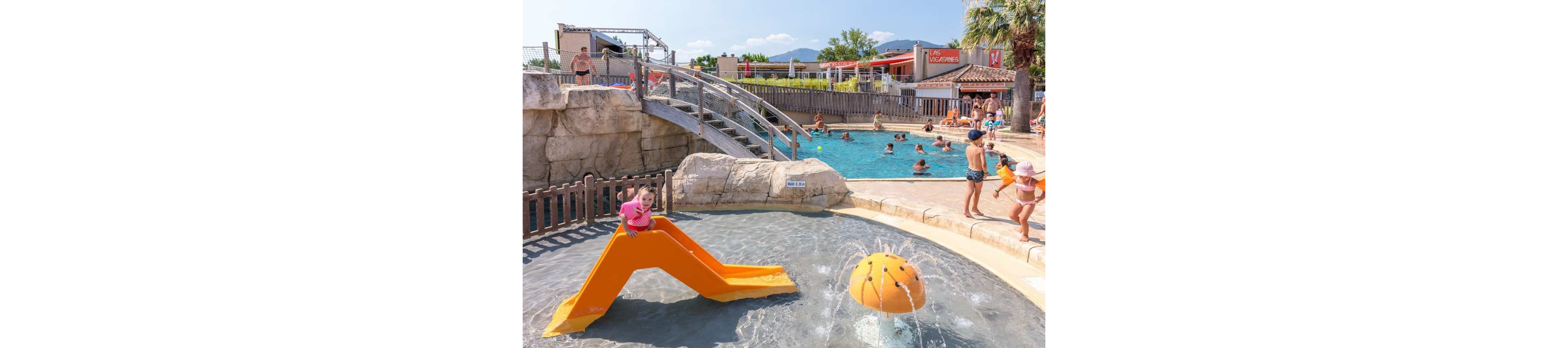 A young child plays on a small orange slide in a shallow splash pool at an outdoor water park. Nearby, a playful water feature sprays water while families swim and relax in a larger pool in the background. The setting includes stone-style structures, a wooden bridge, and palm trees under a bright blue sky. People of various ages enjoy the warm weather and resort-like atmosphere.