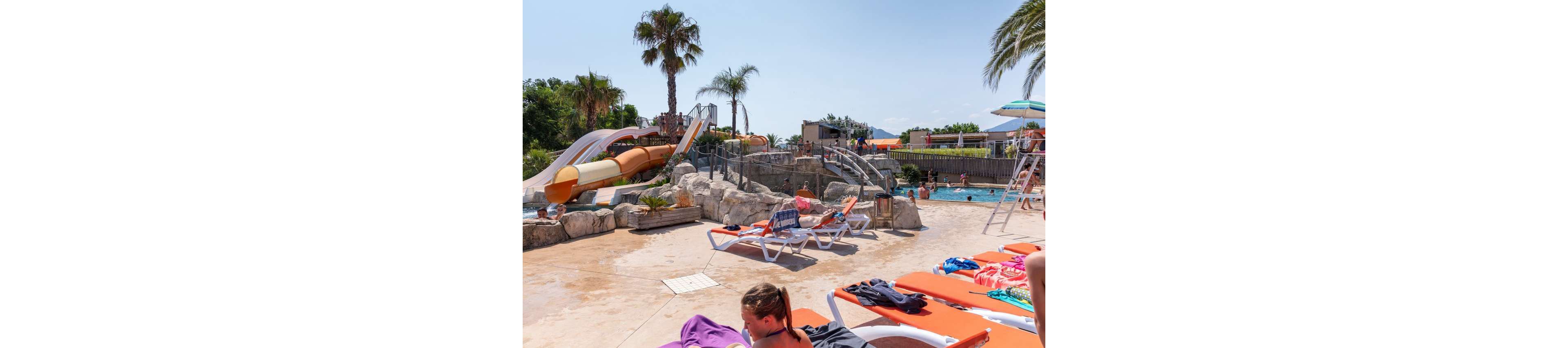 An outdoor water park features colorful tube slides built into rocky landscaping beside a swimming pool. People relax on orange sun loungers while others swim and play in the pool. Palm trees and clear blue sky create a warm, tropical holiday atmosphere. The scene suggests a lively summer day with families enjoying leisure time together.