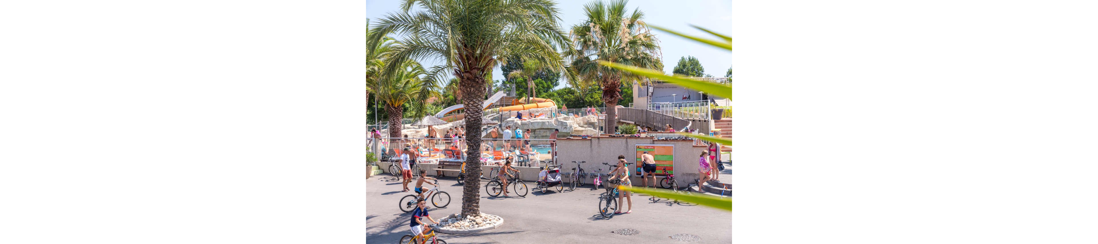 An outdoor playground features a small toy train ride set on a dry, sandy surface. In the foreground, a blue fish-shaped spring rider stands out against the light ground. A colorful inflatable bouncy castle with cartoon pirate characters is visible in the background. Palm trees, a sports court, and a few people with bicycles complete the sunny recreational setting.