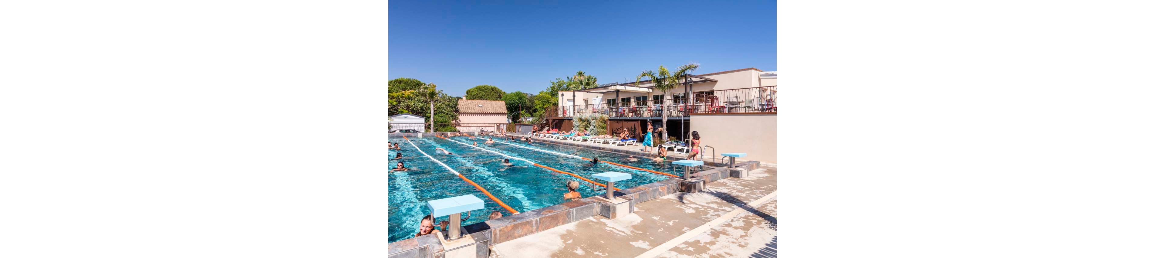 A lively outdoor swimming pool is filled with families and children playing in the water. People relax on sun loungers around the pool while others swim, wade, and play with inflatable toys. A small bridge crosses over part of the pool, adding visual interest to the scene. In the background, low buildings, palm trees, and distant mountains suggest a warm holiday resort atmosphere.