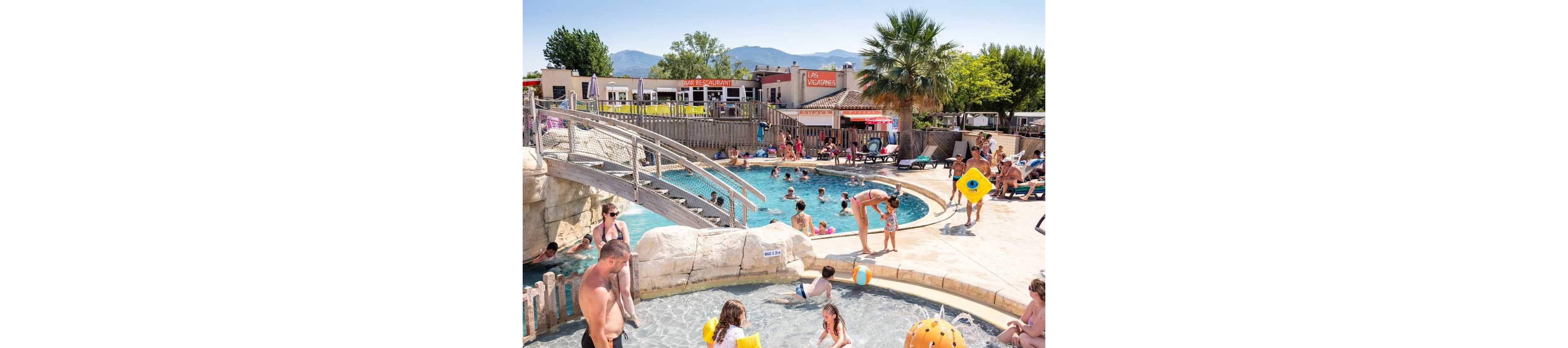 A long rectangular outdoor swimming pool is shown in a sunny resort setting. The pool has lane markings and clear blue water, surrounded by numerous white and beige sun loungers. In the background, there is a covered pool structure and palm trees adding a tropical atmosphere. The sky is clear and blue, suggesting a warm, relaxing vacation environment.