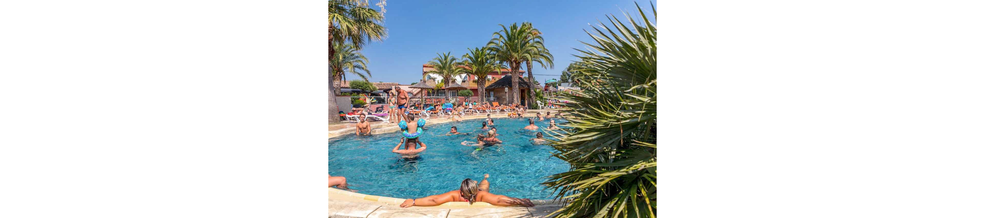 A busy outdoor swimming pool is filled with people enjoying a sunny day. Adults and children play in the water while others relax on the pool edge and surrounding sun loungers. Palm trees and resort buildings frame the scene, creating a tropical holiday atmosphere. The clear blue sky and bright sunlight emphasize a warm, cheerful vacation setting.