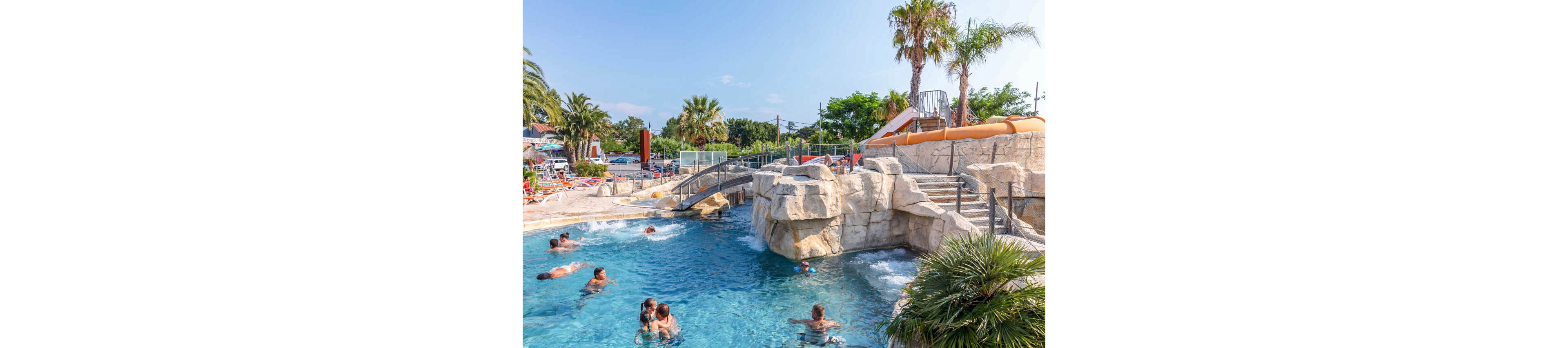 Several people swim and relax in a large outdoor resort pool surrounded by artificial rock formations. A small waterfall cascades into the blue water near a bridge that crosses the pool. In the background, an orange waterslide curves down beside palm trees and sun loungers. The sunny weather and clear sky create a bright, cheerful vacation atmosphere.