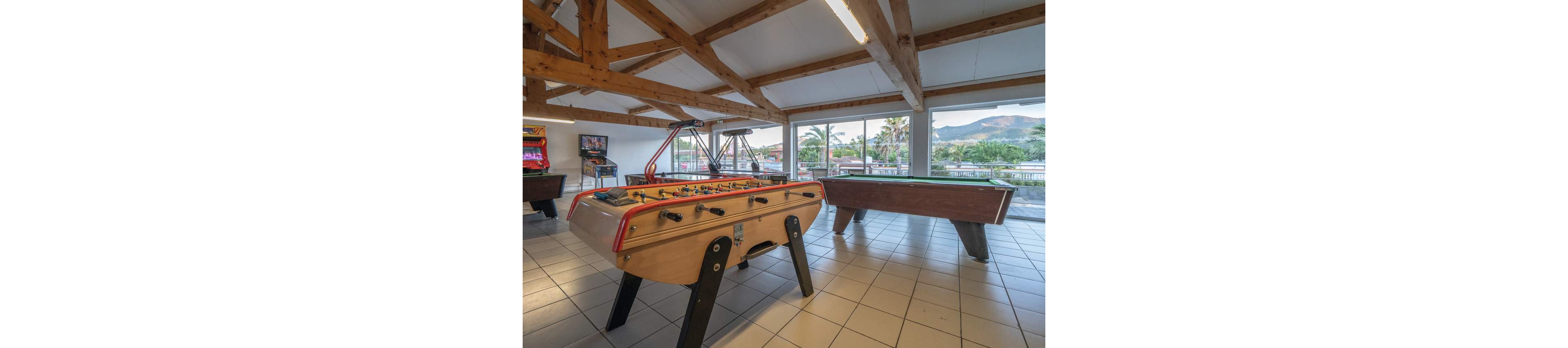 A bright recreation room features a foosball table in the foreground and a pool table nearby. Large windows reveal an outdoor view with palm trees and distant mountains. The interior has tiled floors, exposed wooden beams, and several arcade machines along the walls. The space appears ready for leisure activities and social gatherings.