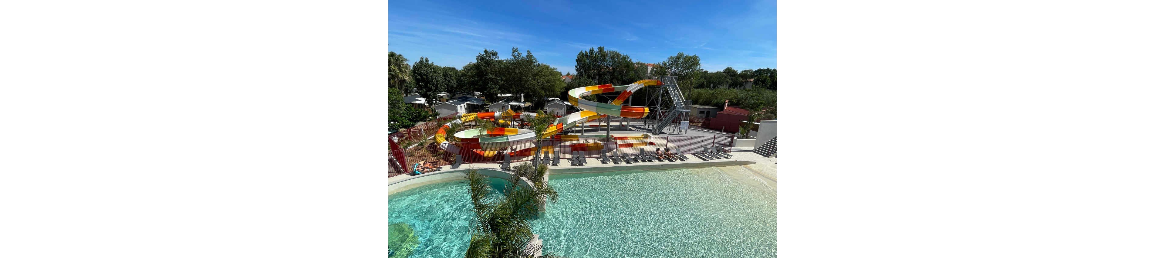 An outdoor water park features large winding water slides in bright yellow, orange, and green colors. The slides empty into a pool area bordered by lounge chairs and umbrellas where people are sunbathing. Clear blue sky and surrounding trees create a relaxed vacation atmosphere. A shallow pool with gentle waves and a palm tree in the foreground adds to the tropical resort feel.