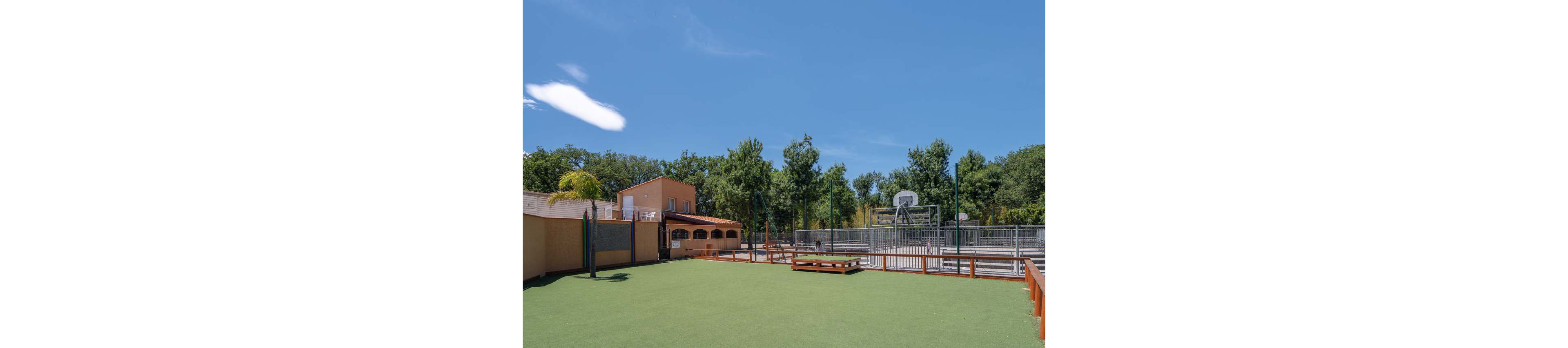 An empty outdoor sports area with artificial grass is shown under a clear blue sky. A fenced basketball court with metal bleachers stands to the right side of the image. On the left, a peach-colored building with arched windows and a terrace borders the court. Tall green trees surround the facility, creating a calm and sunny recreational setting.
