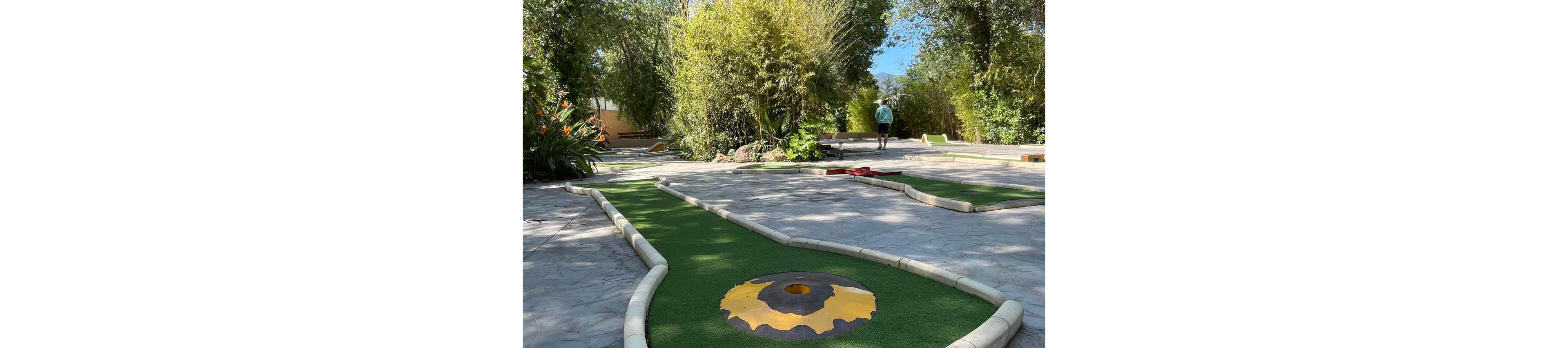 A brightly lit mini golf course winds through a landscaped outdoor area. Artificial green turf is bordered by low white curbs, leading to a large circular hole with a yellow and brown design. Lush trees and bamboo create a shaded, natural backdrop around the course. In the distance, a person walks along the path, adding a sense of scale and leisurely activity.