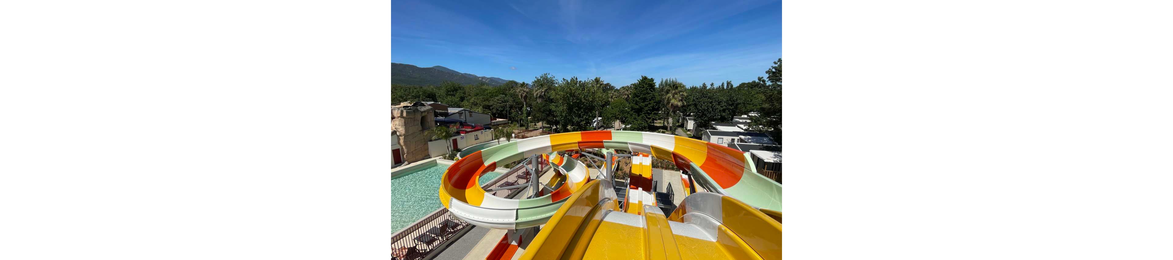 A large, colorful water slide twists above an outdoor pool area at a resort or water park. The slide features bright yellow, orange, green, and white sections curving around a central support structure. Below, lounge chairs and pathways surround the pool, with trees and cabins or buildings in the background. Clear blue sky and distant hills suggest a warm, sunny day ideal for outdoor recreation.