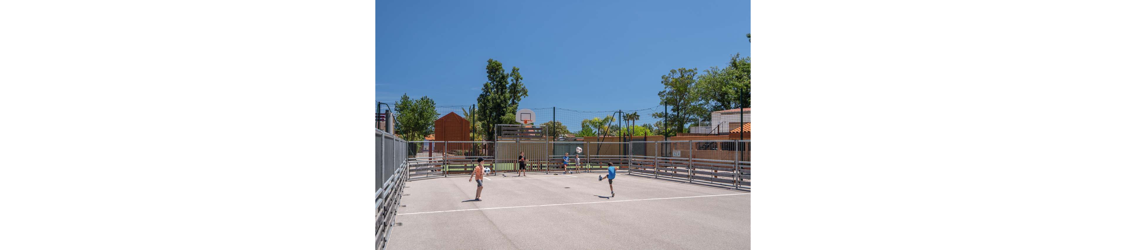A group of children play soccer on a fenced outdoor sports court under a clear blue sky. The court features a central goal area and a basketball hoop at one end. Trees, playground equipment, and low buildings surround the court, suggesting a school or community recreation area. The bright daylight and open space create an energetic, summery atmosphere.