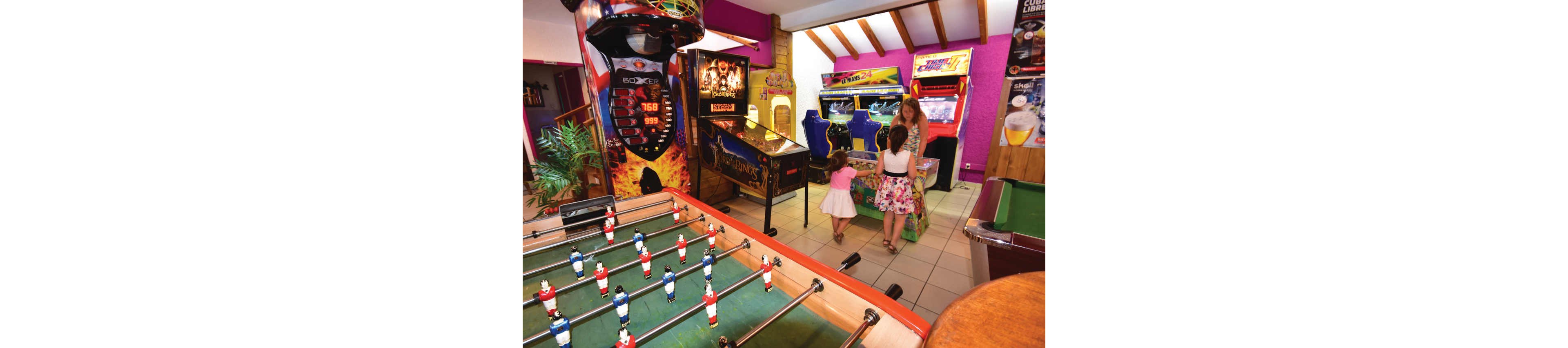 A brightly lit arcade room features several classic arcade machines and a foosball table in the foreground. A woman stands beside two young girls who are playing at one of the machines. The setting has vivid pink and yellow walls, tiled flooring, and exposed wooden ceiling beams, creating a lively atmosphere. The scene conveys family fun, leisure, and entertainment in an indoor game space.