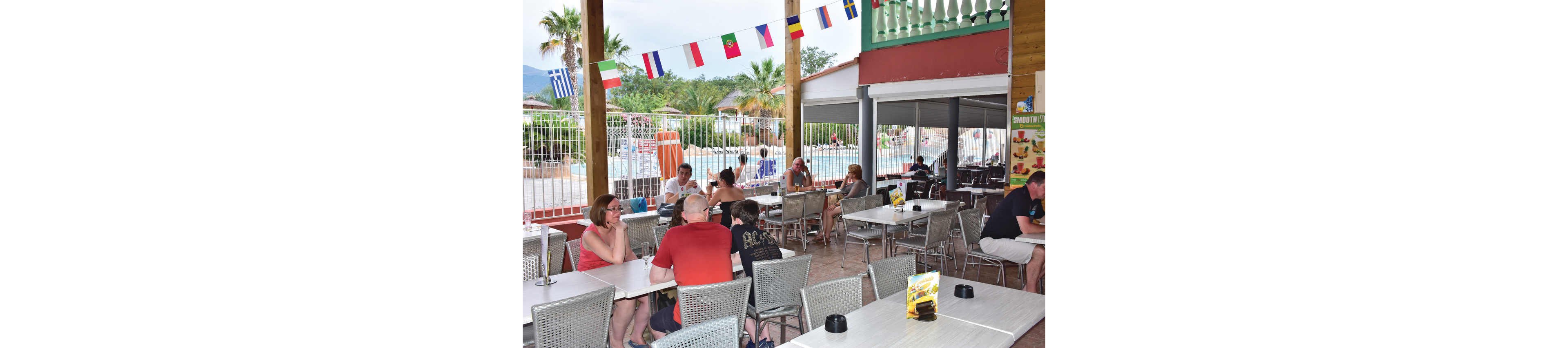 An open-air café terrace overlooks a swimming pool where people relax on a sunny day. Several adults and children sit at metal tables, talking, drinking, and enjoying the casual atmosphere. Colorful national flags hang from the wooden beams above the seating area, adding to the vacation setting. In the background, palm trees, sun loungers, and a fenced pool area suggest a resort or holiday park environment.