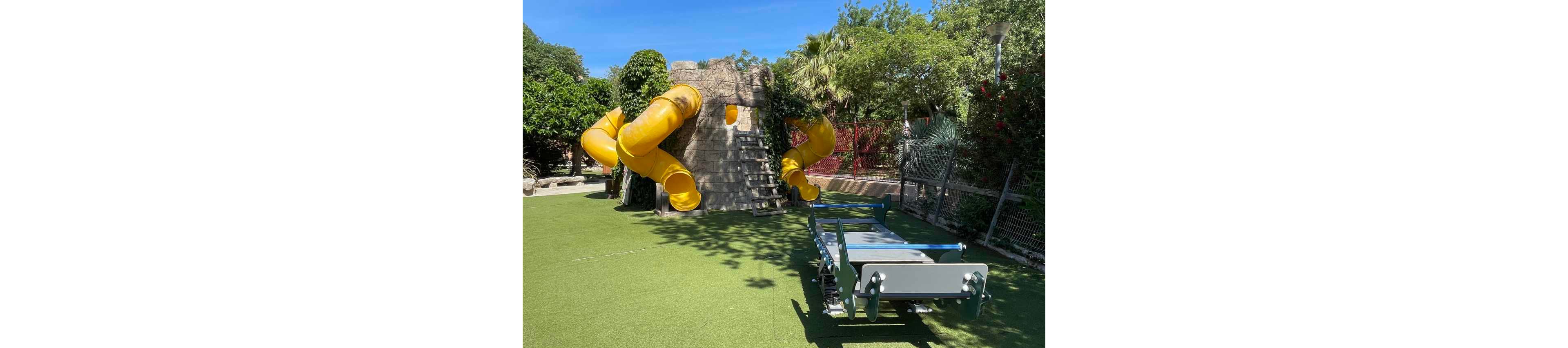 An outdoor playground features a large climbing structure with multiple bright yellow tube slides. The structure is set on a wide area of artificial grass under a clear blue sky. A gray and green playground platform or seesaw sits in the foreground, casting shadows on the turf. Lush green trees, shrubs, and fencing surround the play area, creating a safe and enclosed environment.